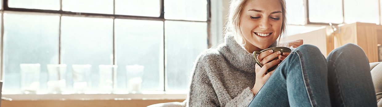 Lächelnde Frau mit Tasse auf dem Sofa – Moment der Entspannung bei PMS-Symptomen